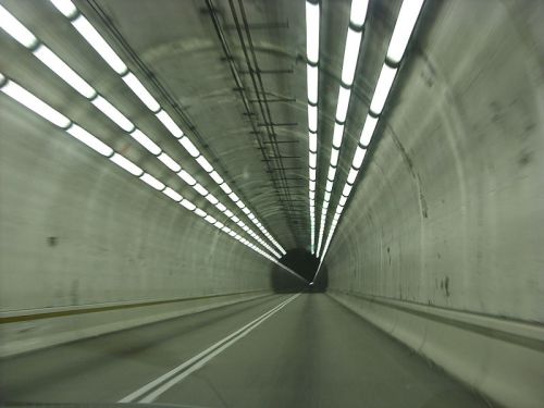 A view inside a tunnel with bright overhead lights and a dark exit ahead, featuring a smooth road surface.