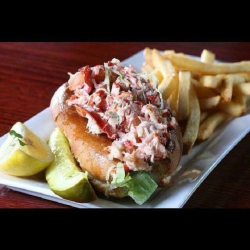 A lobster roll topped with lobster salad, served with fries and lemon wedges on a white plate.