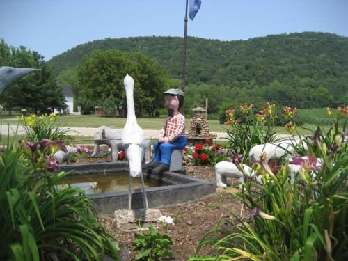 A whimsical garden scene featuring a statue of a man sitting by a pond, surrounded by colorful flowers and greenery.