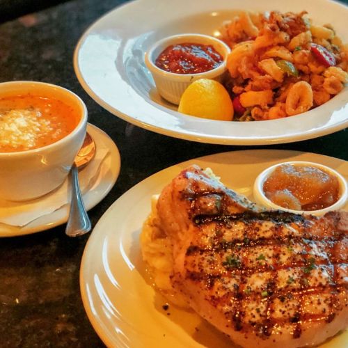 A plate with grilled pork chop, mashed potatoes, and apple sauce, alongside a bowl of soup and a dish of fried calamari.