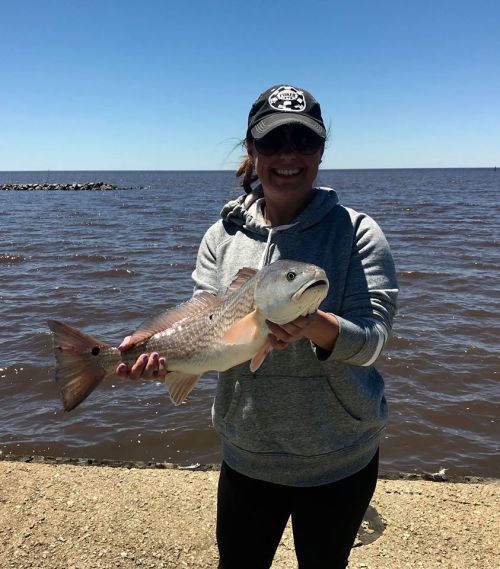 A person in a gray hoodie and cap holds a large fish by the water on a sunny day.