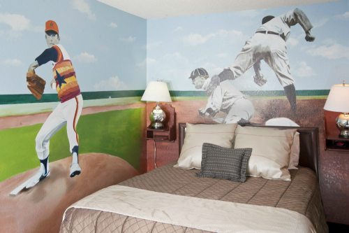 A bedroom featuring a baseball-themed mural of a pitcher and a player sliding into base, with a cozy bed and lamps.