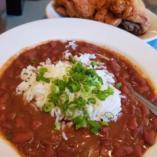 A bowl of red beans and rice topped with green onions, with a side of fried chicken in the background.