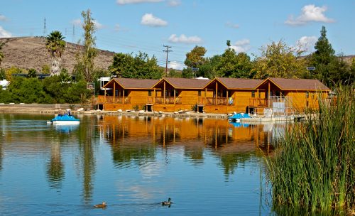 A serene lake scene featuring wooden cabins reflecting on the water, surrounded by greenery and a clear blue sky.