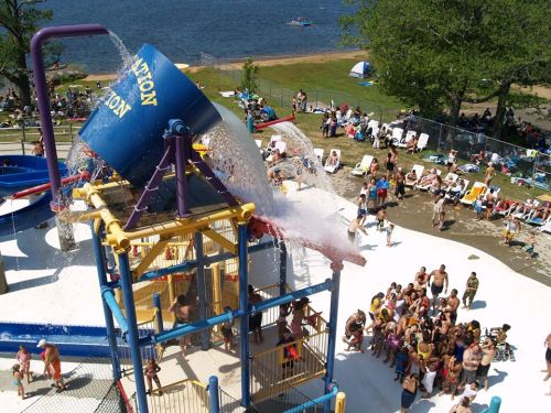 A water park with a large splash bucket pouring water over excited visitors below, surrounded by sunbathers and a lake.