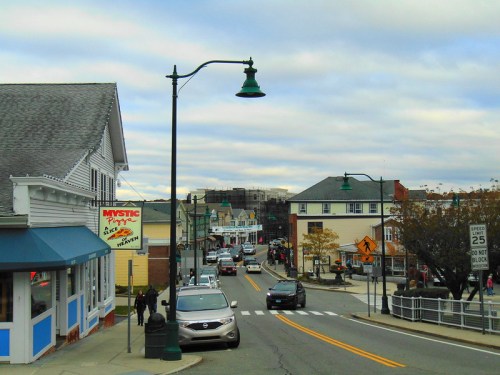 A street view of a quaint town with shops, cars, and a cloudy sky, showcasing a charming small-town atmosphere.