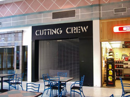 A closed hair salon named "Cutting Crew" with a black shutter, located in a mall with empty tables and chairs nearby.