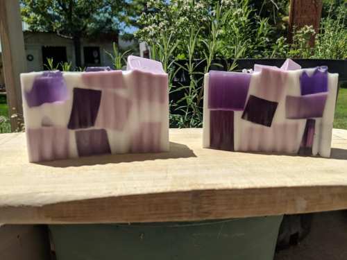 Two colorful handmade soaps with purple and white geometric patterns, displayed on a wooden surface outdoors.