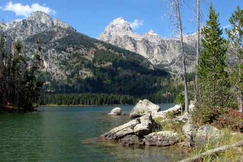 A serene lake surrounded by mountains, with rocky shores and trees under a clear blue sky.