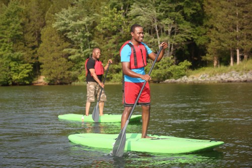 Two men paddleboarding on a calm lake surrounded by trees, wearing life jackets and enjoying a sunny day.