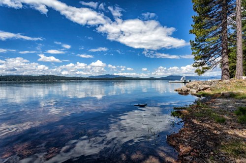 A serene lake scene with calm waters, blue skies, fluffy clouds, and trees along the shore.