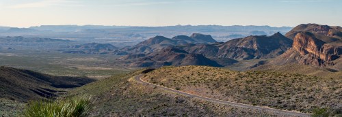 A panoramic view of rugged mountains and valleys under a clear sky, with a winding road in the foreground.