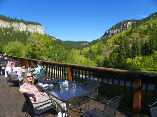 A woman sits at a table on a deck, surrounded by lush green mountains and clear blue skies.