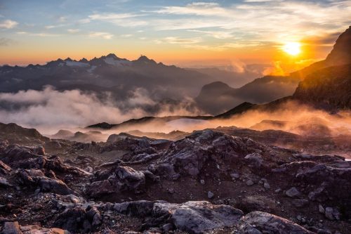 A breathtaking sunset over rugged mountains, with clouds and mist swirling around rocky terrain.