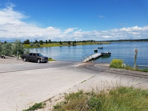 A truck parked near a boat ramp by a calm lake, with boats and a dock in the background under a blue sky.