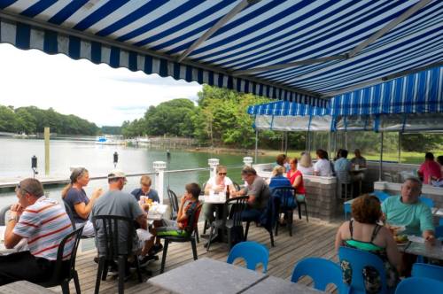 A waterfront dining area with people enjoying meals under a blue and white striped awning. Trees and water in the background.