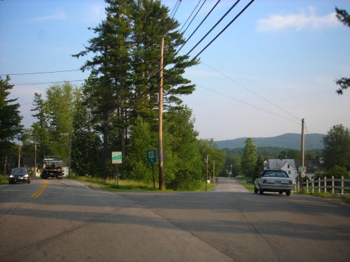 A rural intersection with trees, power lines, and a sign for Route 138, under a clear blue sky.
