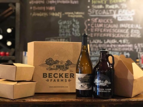 A wooden table displays bottles and a growler from Becker Farms Brewing Co., alongside takeout boxes and a chalkboard menu.
