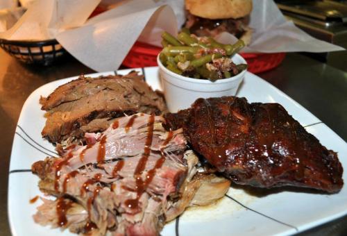 A plate of sliced barbecue meat with sauce, accompanied by green beans in a cup and a sandwich in the background.