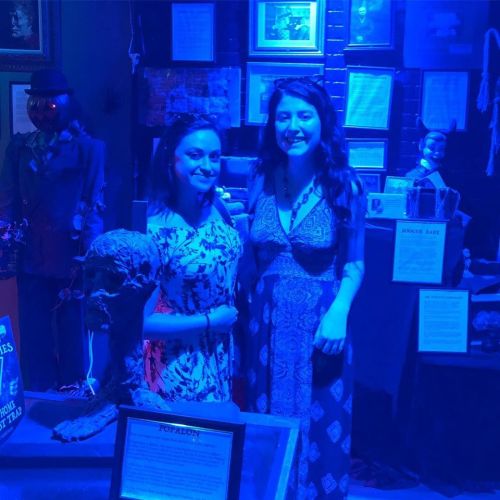 Two women pose together in a dimly lit room with blue lighting, surrounded by eerie displays and artifacts.