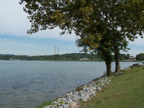A serene lakeside view with trees, rocky shoreline, and distant smokestacks under a clear blue sky.