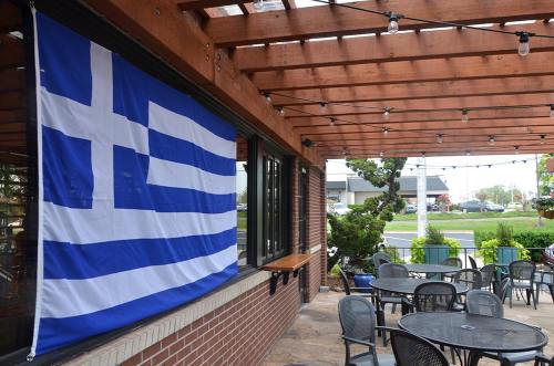 A Greek flag hangs on a brick wall beside an outdoor dining area with empty tables and string lights overhead.