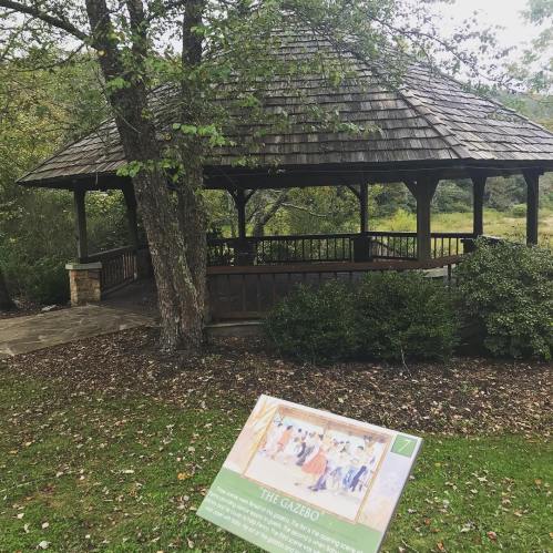 A wooden gazebo surrounded by greenery, with an informational sign in the foreground.