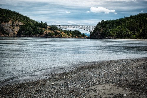 A serene river scene with a bridge spanning across, surrounded by lush green hills under a cloudy sky.