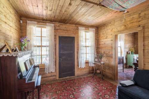 Cozy wooden room with a piano, floral rug, and lace curtains, featuring a warm, rustic atmosphere.
