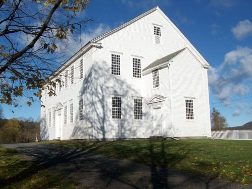 A large white building with multiple windows, set against a blue sky and surrounded by green grass and trees.