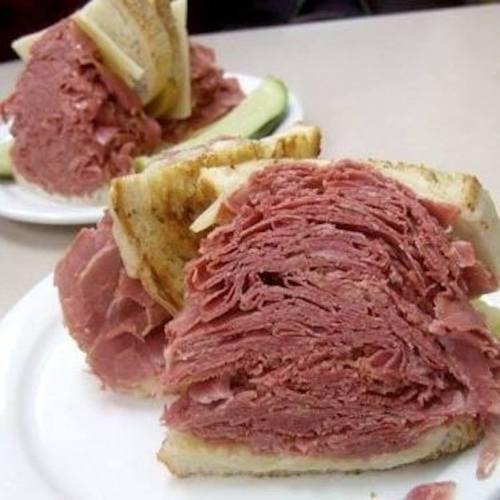 A close-up of a stacked pastrami sandwich on white bread, served with pickles on the side.
