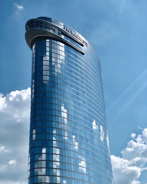 A tall, modern glass skyscraper reflecting clouds against a blue sky. The building features a curved top.