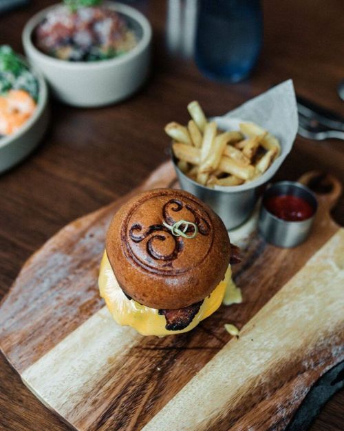 A gourmet burger with cheese on a wooden board, accompanied by a small metal cup of fries and ketchup.