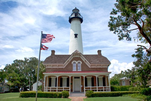 A tall lighthouse stands behind a historic house, with American and state flags waving in the foreground under a blue sky.