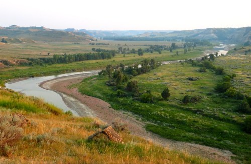 A winding river flows through a lush green valley, surrounded by rolling hills and distant mountains under a soft sky.