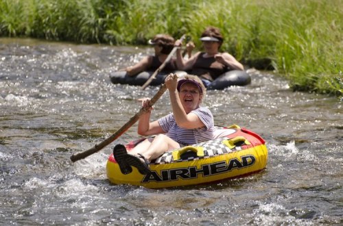 A woman joyfully paddles a red inflatable tube in a river, with two others floating in the background.