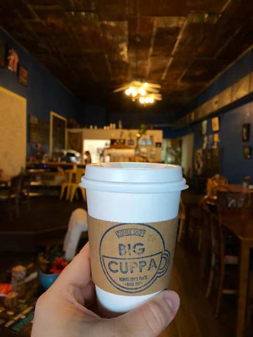 A hand holds a coffee cup with "Big Cuppa" logo in a cozy coffee shop with blue walls and wooden furniture.