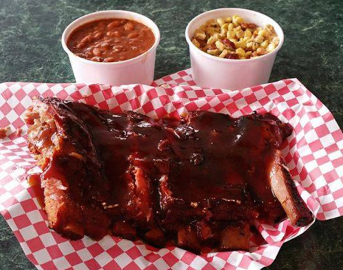 A plate of barbecue ribs with a glossy sauce, accompanied by cups of baked beans and corn salad on a checkered paper.
