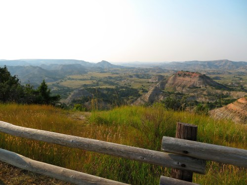 A scenic view of rolling hills and valleys under a hazy sky, framed by a wooden fence and tall grass.