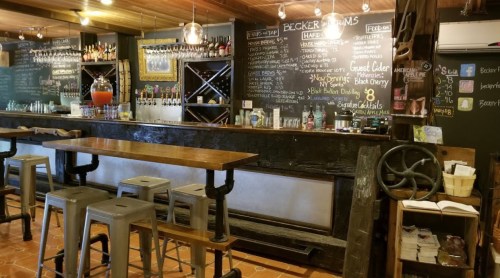 A rustic bar with wooden tables, metal stools, and a chalkboard menu displaying drinks and specials.