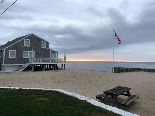 A beach scene at sunset with a gray house, American flag, and picnic table on sandy shore under cloudy skies.