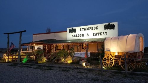 A rustic saloon and eatery with a wagon outside, illuminated by lights at dusk, featuring a sign that reads "Stampede Saloon & Eatery."