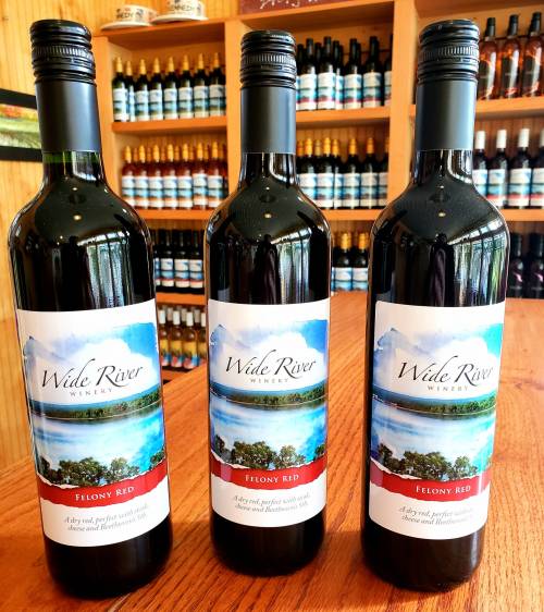 Three bottles of Wide River Winery Felony Red wine on a wooden table, with a backdrop of wine bottles.