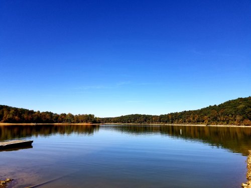 A calm lake reflects a clear blue sky, surrounded by trees with autumn foliage.