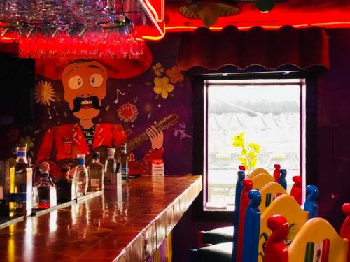 Colorful bar scene featuring a mural of a musician, bottles on the counter, and vibrant chairs under red lighting.