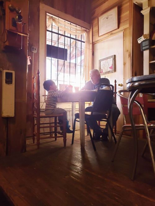 A child and an adult sit at a table in a cozy, rustic room, engaged in conversation by a window.