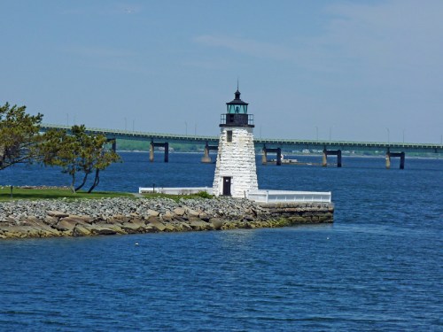 A white lighthouse stands on a rocky shore, with a bridge in the background and calm blue waters surrounding it.