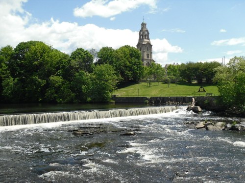 A serene river with a small waterfall, lush green trees, and a historic building in the background under a blue sky.