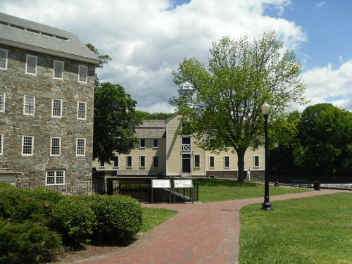 Historic buildings with stone and wooden facades, surrounded by green grass and trees under a partly cloudy sky.