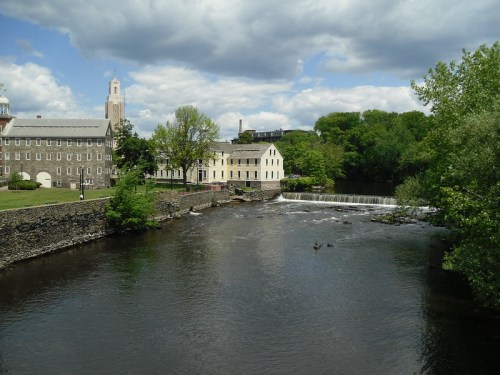 A serene river scene with a waterfall, green trees, and historic buildings under a partly cloudy sky.